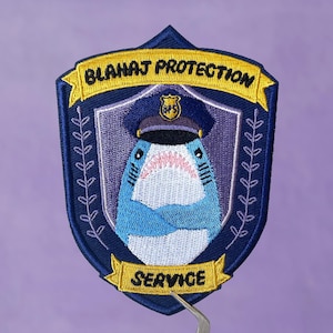 May include: An embroidered patch featuring a shark wearing a police hat, with the text "Blah blah Protection Service" in blue and yellow.