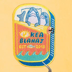 Puede incluir: Parche bordado con dos tiburones azules en una lata roja y amarilla con el texto "KEA BLAHAJ EST 2010" y un sol amarillo con un ojo.