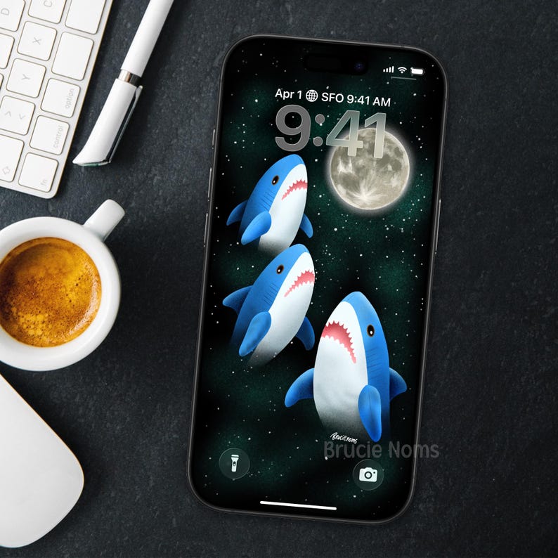 May include: A smartphone displays a whimsical wallpaper featuring three cartoon sharks with blue bodies and white bellies, set against a starry green background with a full moon. The time is 9:41 AM, and the text "Brucie Noms" is visible.