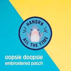 May include: A blue and white embroidered patch with a shark and the text "Hangry All The Time".