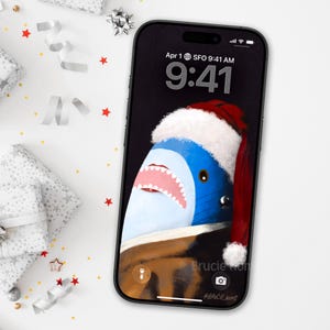 May include: A black smartphone displays a shark wearing a Santa hat on its screen. The shark is blue and white with a red hat and a white trim. The screen shows the time as 9:41 AM, with the date April 1st. The artist's name is visible.