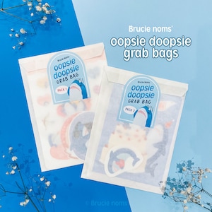 May include: Two white envelopes with blue and white graphics. The envelopes are labeled "oopsie doopsie grab bag" and have a shark illustration. The text "Brucie noms'" is at the top of the image.