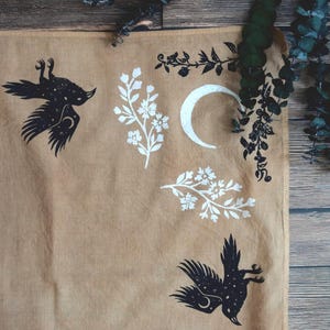 May include: A tan fabric with black and white celestial and floral designs. The illustrations include a crescent moon, birds with star patterns, and floral branches. The background is a light brown color.