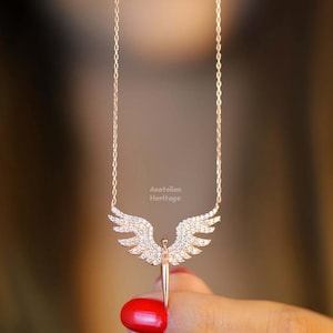 May include: A rose gold necklace featuring a sparkling angel pendant. The angel has wings and a body made of small, clear stones. The necklace is on a chain with a delicate design.
