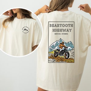 May include: A cream colored t-shirt with a graphic on the back that says "Beartooth Highway Montana | Wyoming" and features a person riding a dirt bike through a mountain landscape. The front of the shirt has a small graphic of mountains with the text "Motorcycle Roads of the Teton Range".