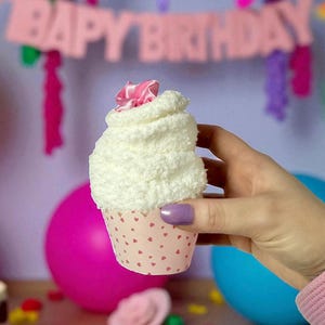 May include: A decorative cupcake made of white fabric with a pink ribbon on top, held in a pink cupcake liner with red hearts. The background includes a pink balloon, a blue balloon, and a banner that says "HAPPY BIRTHDAY".