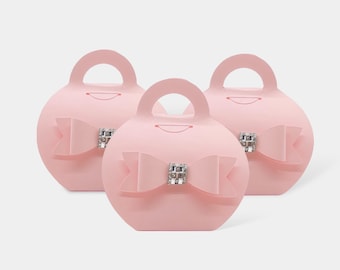May include: Three light pink round gift boxes with integrated handles. Each box features a centered paper bow decoration adorned with a square silver-toned embellishment containing clear crystals at the center of the bow on the front side.
