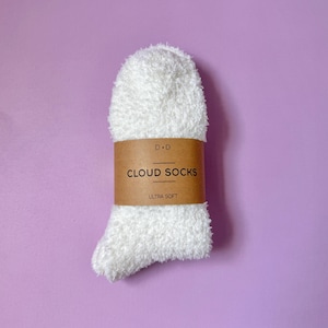 May include: A pair of white cloud socks with a brown label wrapped around the middle. The label reads "CLOUD SOCKS" in a bold font, with "ULTRA SOFT" below. The socks are fluffy and soft, set against a purple background.