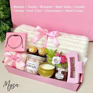 May include: A pink gift box filled with a white towel, bath salts, a candle, chocolates, honey, and a hair claw. The box is decorated with a pink ribbon and flowers. The text "Mysa" is printed on the bottom of the box.