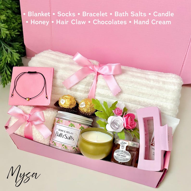 May include: A pink gift box filled with a white towel, bath salts, a candle, chocolates, honey, and a hair claw. The box is decorated with a pink ribbon and flowers. The text "Mysa" is printed on the bottom of the box.