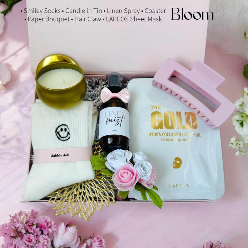 May include: A pink gift box filled with self-care items, including a candle, a bottle of linen spray, a sheet mask, a hair claw, and a pair of socks with a smiley face. The box is decorated with a pink flower bouquet and the word "Bloom" is printed on the top.