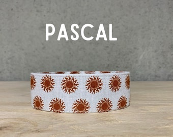May include: A white fabric dog collar with a repeating pattern of orange suns. The collar is labeled "PASCAL".