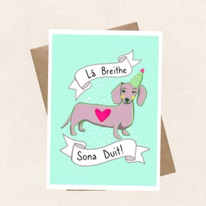 May include: A light blue card featuring a pink dachshund wearing a green party hat and a pink heart. The card reads "Lá Breithe Sona Duit!" which is Irish for "Happy Birthday to You!"