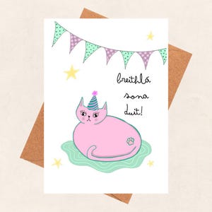 May include: A pink cat wearing a party hat with a green and purple banner above. The text on the card says "breithla sona duit!" which is Irish for "Happy Birthday to you!"