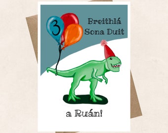 Pode incluir: Um dinossauro verde com um chapéu de festa vermelho e branco segura três balões, um azul, um vermelho e um laranja. O cartão diz "Breithla Sona Duit a Ruan!" que se traduz como "Feliz aniversário!" em irlandês.