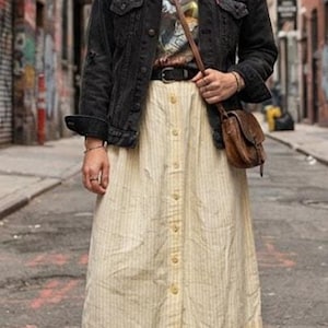May include: A black denim jacket worn over a dark gray graphic t-shirt and a light yellow midi skirt with vertical pinstripes and a front button closure. The outfit includes a brown leather crossbody bag, a black belt, and white high-top sneakers.