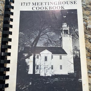 Könnte beinhalten: Ein spiralgebundenes Kochbuch mit dem Titel 1717 MEETINGHOUSE COOKBOOK und einem schwarz-weißen Foto eines Kirchengebäudes auf dem Einband. Der Text West Parish Congregational Church ist unten auf dem weißen Einband in Schwarz gedruckt.