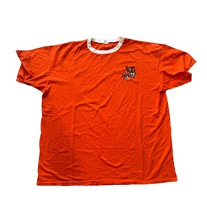 May include: Bright orange t-shirt with a white crew neck and short sleeves. A small graphic of a bear is on the left chest. The shirt is made of a soft material.