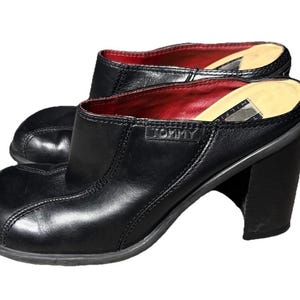 Puede incluir: Un par de mules de cuero negro con tacón de bloque y punta cuadrada. El forro interior es rojo y la palabra TOMMY está grabada en el lateral. Una etiqueta rectangular con el texto TOMMY HILFIGER es visible en la plantilla de color beige.