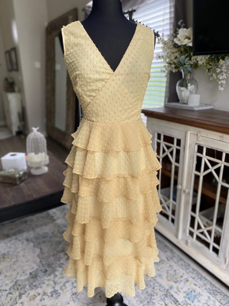 A photo of the sample dress on a mannequin showing the full front of the dress. It has a faux wrap neckline and is sleeveless. The skirt has evenly spaced rufled tiered layers creating a romantic and sassy vibe.