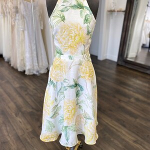 May include: A white linen dress with a halter neckline and a floral print featuring yellow and green flowers. The dress has a fitted bodice and a flowing skirt.