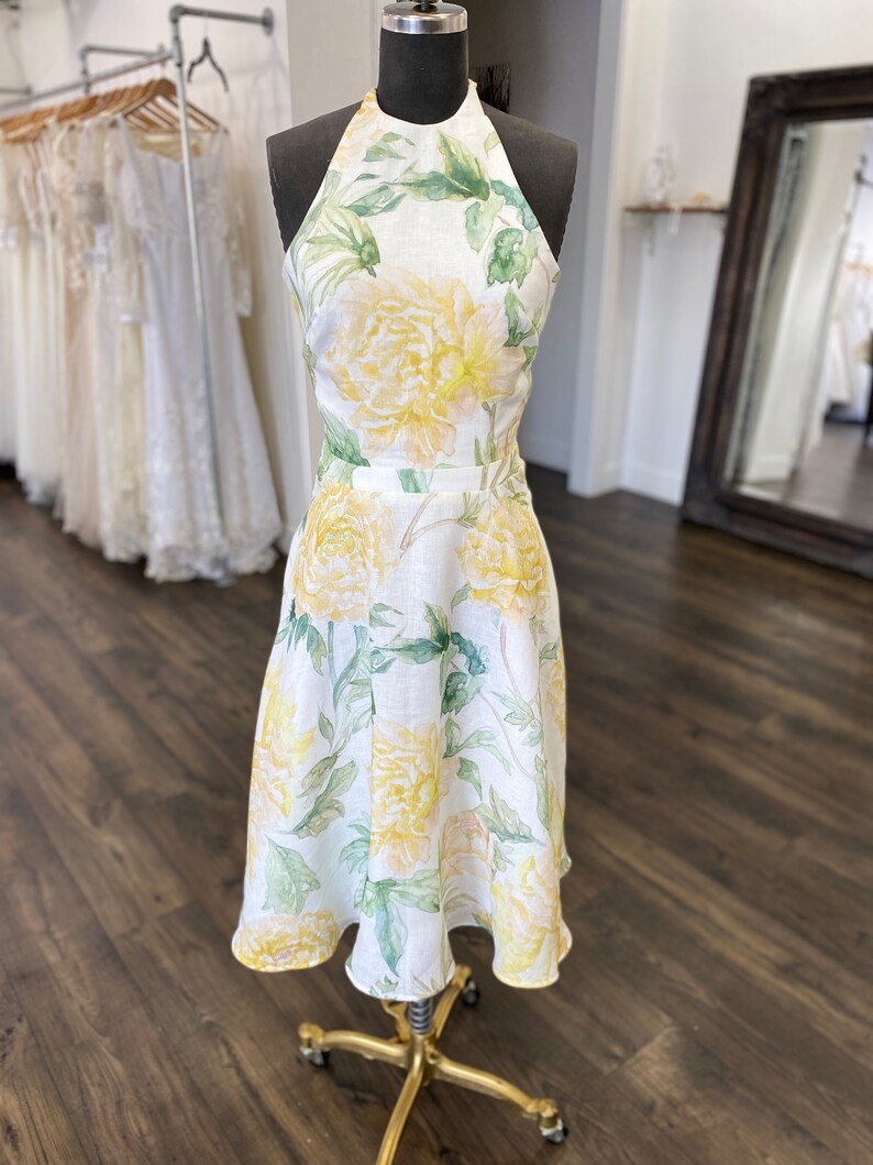 May include: A white linen dress with a halter neckline and a floral print featuring yellow and green flowers. The dress has a fitted bodice and a flowing skirt.