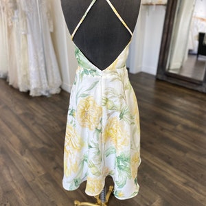 May include: A white midi dress with a yellow floral print and a criss-cross back strap. The dress is on a mannequin and has a fitted bodice and a flowy skirt.