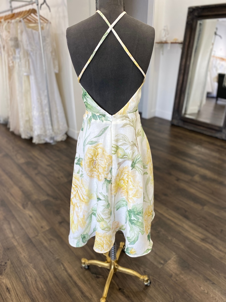 May include: A white midi dress with a yellow floral print and a criss-cross back strap. The dress is on a mannequin and has a fitted bodice and a flowy skirt.