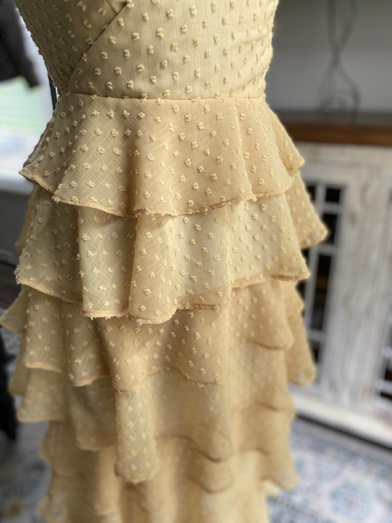 A photo of the sample dress on a mannequin showing a close up on the skirt layers. The fabric used is a clip spot with a chiffon base. It is a beautiful sunflower yellow color and has little rectangular spots. The rectangular spots are raised and have texture like a pile. The skirt layers are all the same length and have a nice flow to bottom while not having any gathers at the top.