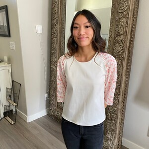 A photo of the sample top on a young woman showing the full front of the top. It has an all white center with a contrasting ralgan sleeve detail. The raglan sleeves are made from a white clip spot fabric with a dainty red print. The top has a high scoop neckline and an A-line silhouette. She wears the top with black skinny jeans.