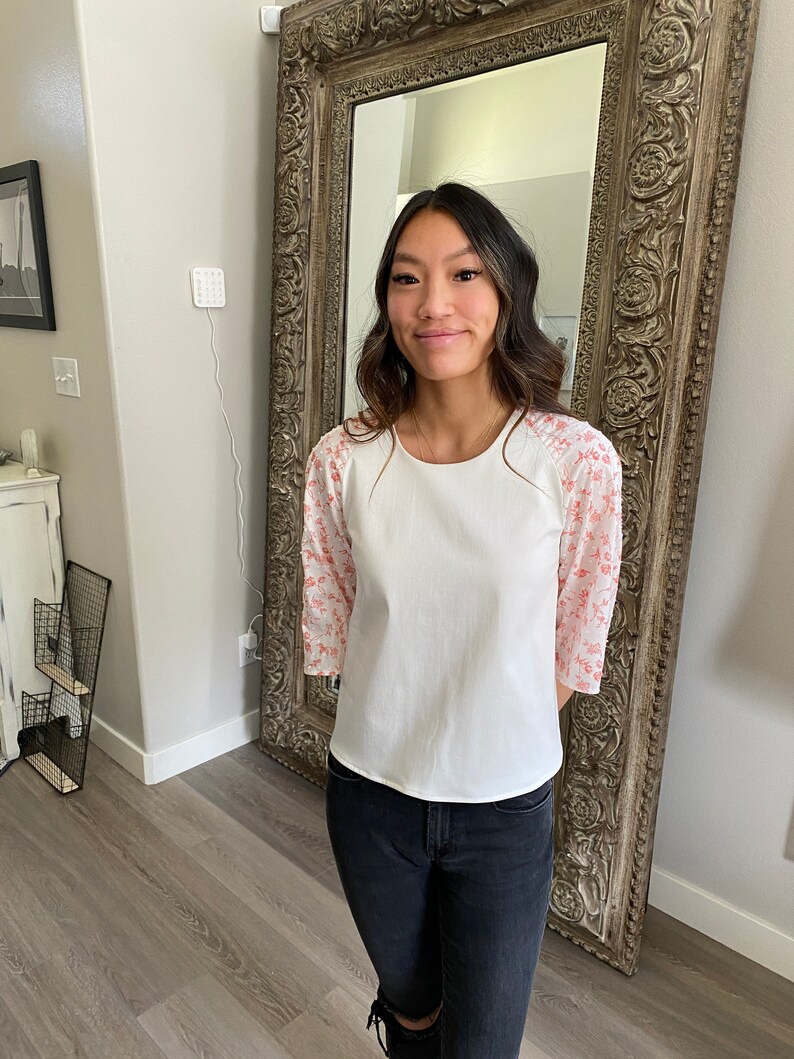 A photo of the sample top on a young woman showing the full front of the top. It has an all white center with a contrasting ralgan sleeve detail. The raglan sleeves are made from a white clip spot fabric with a dainty red print. The top has a high scoop neckline and an A-line silhouette. She wears the top with black skinny jeans.