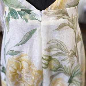 May include: A white linen dress with a floral print featuring yellow flowers and green leaves. The dress has a V-neckline and a zipper closure in the back.