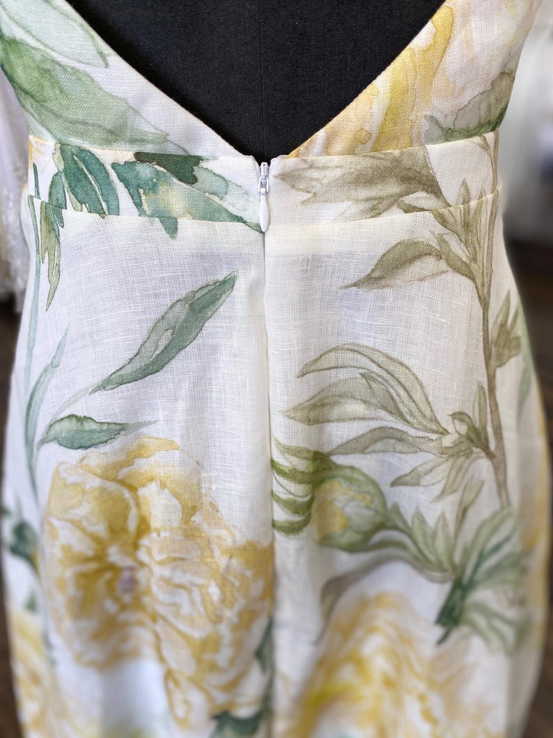 May include: A white linen dress with a floral print featuring yellow flowers and green leaves. The dress has a V-neckline and a zipper closure in the back.