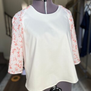 A photo of the sample top on a mannequin showing the full front of the top. It has an all white center with a contrasting ralgan sleeve detail. The raglan sleeves are made from a white clip spot fabric with a floral red print. The top has a high scoop neckline, flared bell raglan sleeves, and an A-line silhouette.