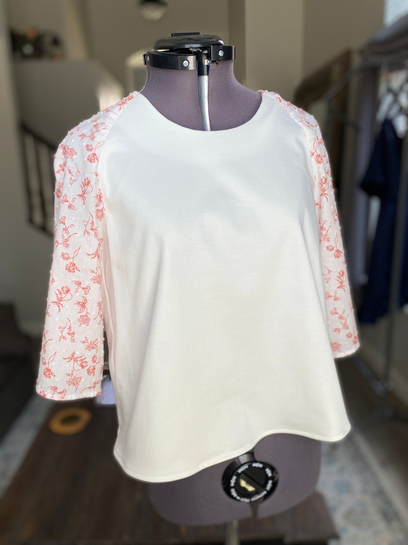 A photo of the sample top on a mannequin showing the full front of the top. It has an all white center with a contrasting ralgan sleeve detail. The raglan sleeves are made from a white clip spot fabric with a floral red print. The top has a high scoop neckline, flared bell raglan sleeves, and an A-line silhouette.
