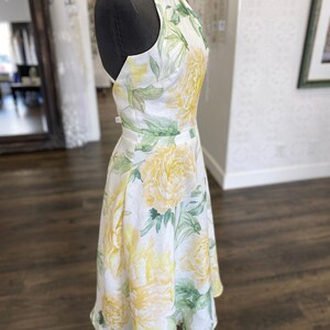 May include: A white linen dress with a yellow and green floral print. The dress has a halter neckline and a fitted bodice. The skirt is A-line and falls to the knee.