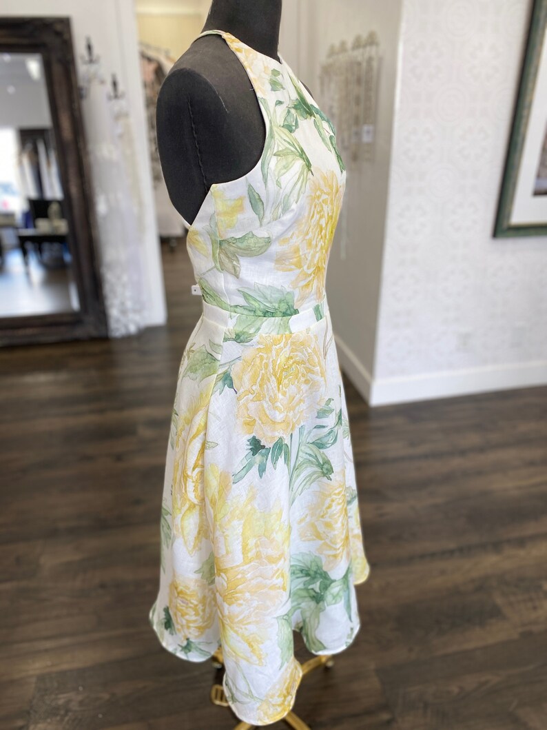 May include: A white linen dress with a yellow and green floral print. The dress has a halter neckline and a fitted bodice. The skirt is A-line and falls to the knee.
