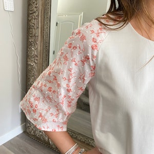 A photo of the sample top focusing on the sleeve. A young woman wears the top and you can see her in the edge of the frame. The top has an all white center with a contrasting ralgan sleeve detail. The raglan sleeves are made from a white clip spot fabric with a dainty floral red print. The raglan sleeve ends just past the elbow.