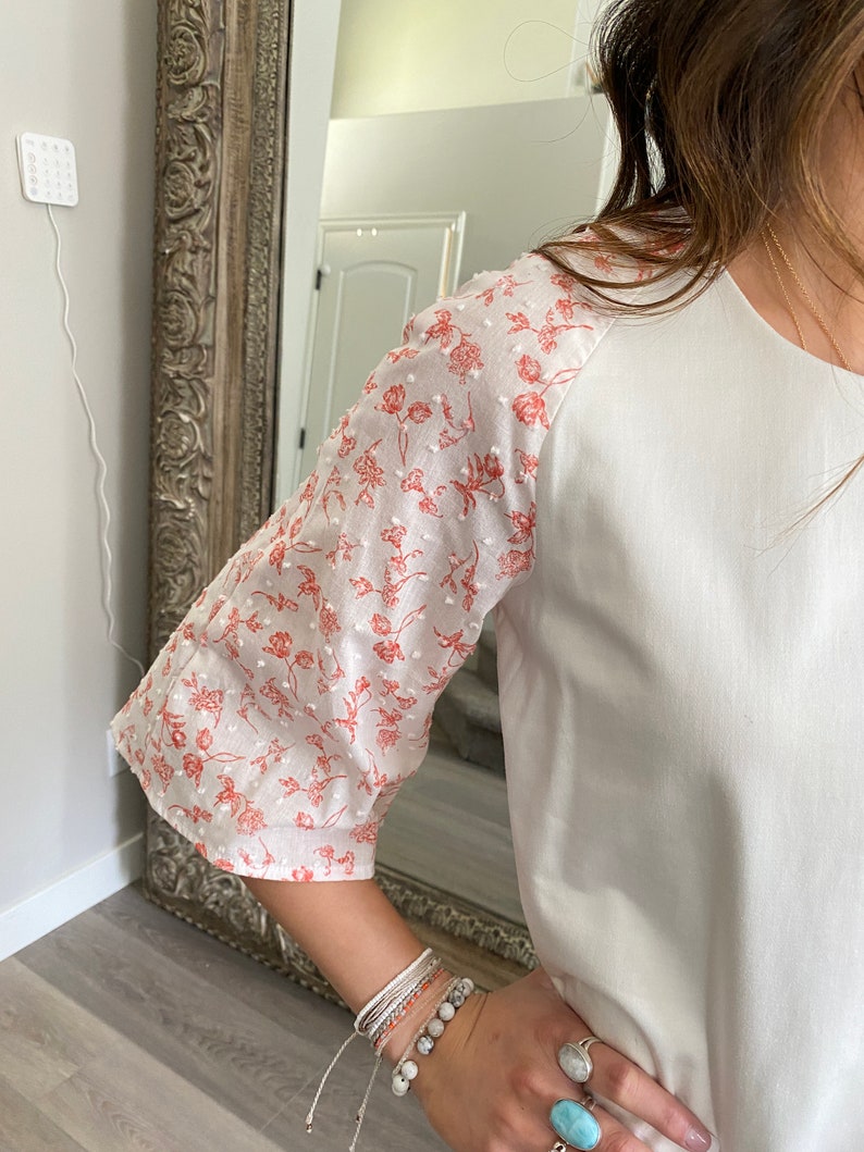 A photo of the sample top focusing on the sleeve. A young woman wears the top and you can see her in the edge of the frame. The top has an all white center with a contrasting ralgan sleeve detail. The raglan sleeves are made from a white clip spot fabric with a dainty floral red print. The raglan sleeve ends just past the elbow.