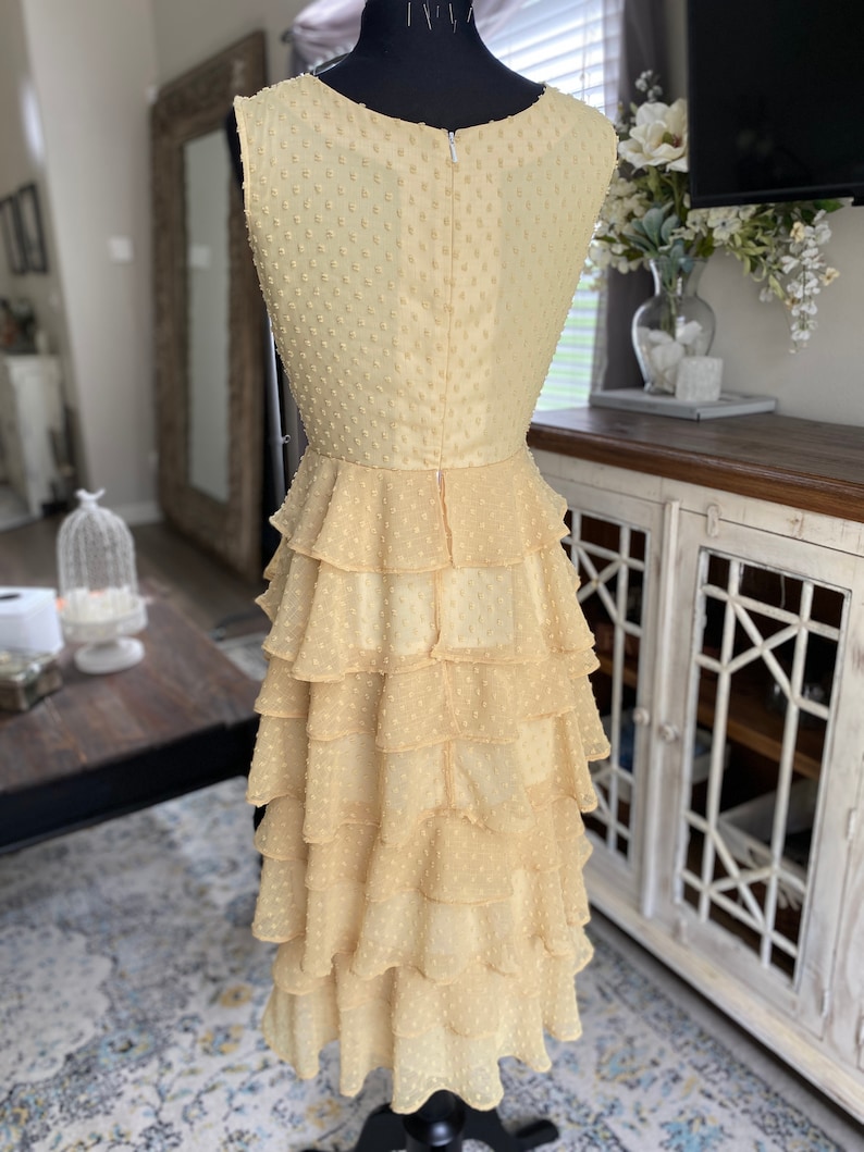 A photo of the sample dress on a mannequin showing the full back of the dress. The back is full coverage with a high scoop neckline and sleeveless shoulder. There is an invisable zipper down the back of the dress. The skirt has ruffled tiers of chiffon layers and is a midi length.