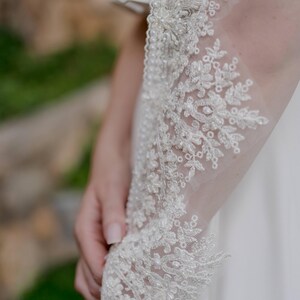 May include: Close-up of a white wedding dress sleeve with intricate floral lace detailing. The lace is embellished with small, sparkling beads. The sleeve is made of a sheer, delicate fabric.