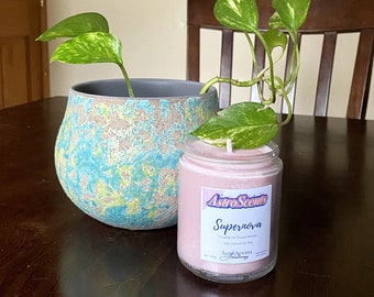May include: A pink candle with a white label that says "AstroScents" and "Supernova" in a clear glass jar. The candle is sitting on a wooden table next to a potted plant with green leaves.