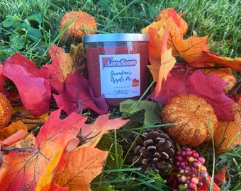 May include: A candle jar with a silver lid and a red label that says "AstroScents Grandma's Apple Pie". The candle is surrounded by fall-colored leaves, a pine cone, and small orange pumpkins.