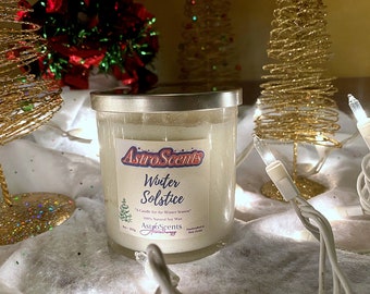 May include: A white candle with a silver lid and a label that reads "Astro Scents Winter Solstice" and "A Candle for the Winter Season" with a small pine tree graphic. The candle is surrounded by white faux fur and gold wire Christmas trees with white lights.