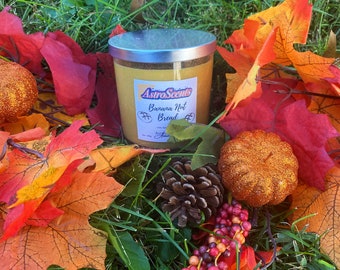 May include: A yellow candle with a silver lid and a label that reads "AstroScents Banana Nut Bread" surrounded by fall foliage, including red and orange leaves, a pine cone, and a small, glittery, orange pumpkin.