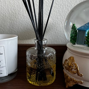 Op de afbeelding: Een heldere glazen fles met een gele vloeistof en zwarte diffuserstokjes. De fles heeft een getextureerd oppervlak en staat op een houten oppervlak. De fles staat naast een witte kaars met een zwart label waarop "Replica" staat en een witte sneeuwbol met een blauw dak.