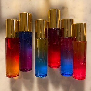 Puede incluir: Seis botellas de vidrio con rodillo y tapas doradas, con líquido de colores en su interior. Las botellas están dispuestas en fila sobre una superficie blanca.