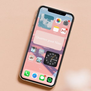 K&ouml;nnte beinhalten: Ein goldenes iPhone mit rosa und blauem Hintergrund. Der Bildschirm des Telefons zeigt ein Uhr-Widget mit der Zeit "Quarter past Nine" und mehrere App-Symbole, darunter Instagram, TikTok, Snapchat, Pinterest und einen Kompass. Das Telefon befindet sich auf einer rosa Oberfl&auml;che.