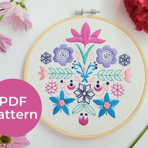 May include: A colourful embroidery hoop with a floral design. The design features pink, blue, green, and purple flowers, leaves, and snowflakes. The hoop is made of wood and has a white fabric background. The text "PDF Pattern" is visible in the bottom left corner of the image.