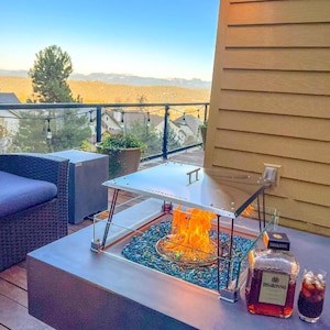 Puede incluir: Un pozo de fuego de acero inoxidable con una tapa de vidrio y un inserto de pozo de fuego de vidrio azul. El pozo de fuego está en una terraza con vista a un bosque y montañas. Hay una botella de licor Disaronno y un vaso de líquido oscuro en la mesa junto al pozo de fuego.